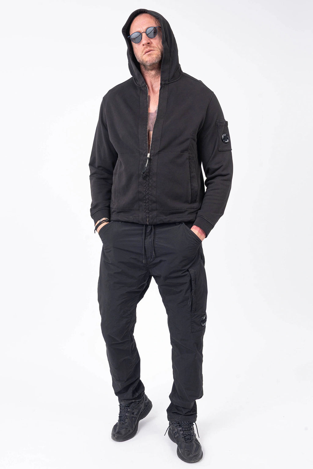 C.P. Company, Mens Sweatshirt, Black 999, Full-Zip, Lens Sleeve Pocket, Hooded, Side Pockets, Ribbed and Cuffs, Cord Adjustable Hem, Snap Closure Sleeve Pocket, Signature C.P. Company Lens Detail, Chester Perry, British Saylor, Massimo Osti, Garment Dyeing Techniques, Rigards Sunglasses and C.P. Company Nylon Pants, Full Body Front View Hooded. 
