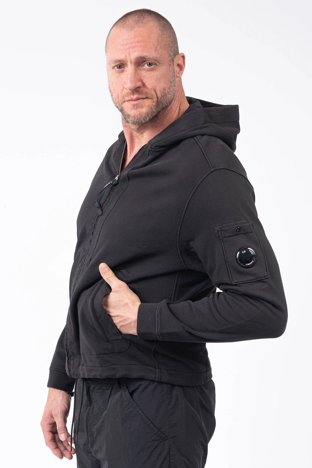 C.P. Company, Mens Sweatshirt, Black 999, Full-Zip, Lens Sleeve Pocket, Hooded, Side Pockets, Ribbed and Cuffs, Cord Adjustable Hem, Snap Closure Sleeve Pocket, Signature C.P. Company Lens Detail, Chester Perry, British Saylor, Massimo Osti, Garment Dyeing Techniques, Front Portrait Hand in Pocket Side View. 