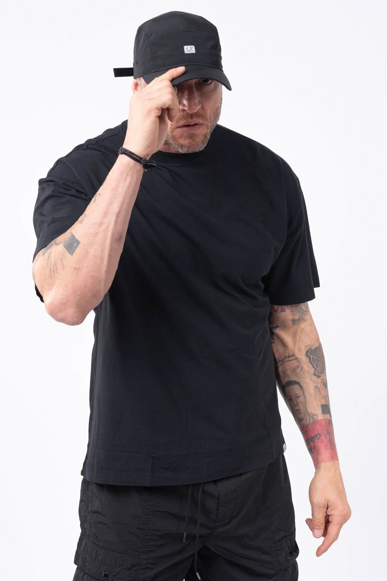 C.P. Company, Mens T-Shirt, Black, Organic Cotton, One Color, Crew Neck, Round Neck, 100% Cotton, Ribbed Neckline and Sleeve Ends, Chester Perry, Massimo Osti, Made in Italy, Front Portrait Wearing a Cap and Pointing to the CP Company Logo. 