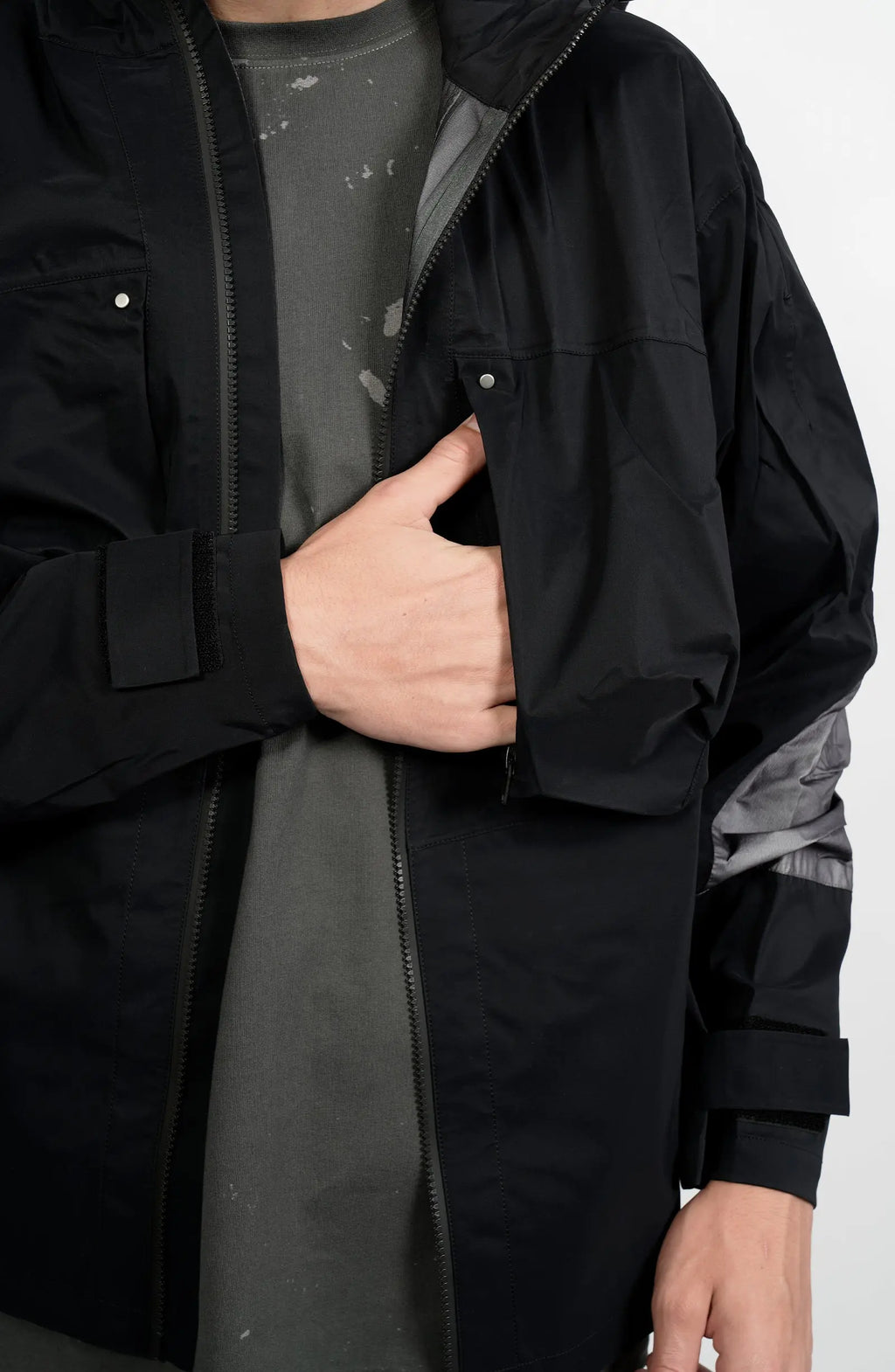 Nilmance, Mens Jacket, Black and Grey, Drawcord Hooded, Velcro, Full Zip, Vislon YKK Zippers, Velcro Adjustsable Cuffs, Bellows Pockets, Side Pockets, Elastic Drawcord, Silver Tone Details, Hand in Front Chest Pocket Close-up. 